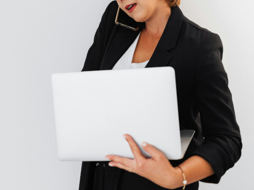 Cropped picture of a woman in a black suit standing against a white wall with an open laptop and a phone stuck between her shoulder and her ear.